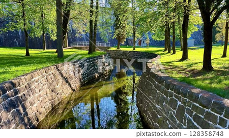 Experience the beauty of a park where a gentle river flows through lush greenery. Surrounded by trees and a stone wall, it invites tranquility Experience the beauty of a park where a gentle river flows through lush greenery. Surrounded by trees and a stone wall, it invites tranquility 128358748