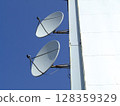 Two white satellite dishes are affixed to the side of a tall structure, facing the clear sky. They are positioned to capture and transmit signals from satellites. 128359329