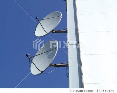 Two white satellite dishes are affixed to the side of a tall structure, facing the clear sky. They are positioned to capture and transmit signals from satellites. 128359329