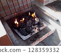 Charcoal glows with flames in a brick fireplace, ready for grilling. The outdoor cooking setup is prepared on a sunny afternoon, inviting a warm atmosphere for gatherings. 128359332