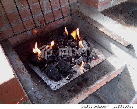 Charcoal glows with flames in a brick fireplace, ready for grilling. The outdoor cooking setup is prepared on a sunny afternoon, inviting a warm atmosphere for gatherings. Charcoal glows with flames in a brick fireplace, ready for grilling. The outdoor cooking setup is prepared on a sunny afternoon, inviting a warm atmosphere for gatherings. 128359332
