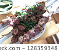 Delicious skewers of grilled marinated meat are garnished with chopped green herbs on a wooden surface. Fresh cucumber slices accompany the dish, creating a savory experience. 128359348