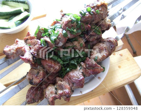 Delicious skewers of grilled marinated meat are garnished with chopped green herbs on a wooden surface. Fresh cucumber slices accompany the dish, creating a savory experience. Delicious skewers of grilled marinated meat are garnished with chopped green herbs on a wooden surface. Fresh cucumber slices accompany the dish, creating a savory experience. 128359348
