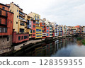 A row of vibrant buildings lines the riverbank in Girona, reflecting their colorful facades in the water. This scene captures the charm and architectural diversity of the area on a cloudy day. 128359355