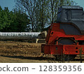 A roller compactor is actively working on a construction site, flattening and compacting soil under a clear blue sky. Nearby, wrapped materials are prepared for use, highlighting ongoing development. 128359356
