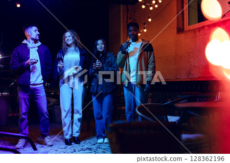 Group of friends enjoying drinks outdoors at night under string lights in a cozy atmosphere Group of friends enjoying drinks outdoors at night under string lights in a cozy atmosphere 128362196