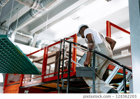 Worker ascending stairs with toolbox in modern industrial workspace during daytime hours 128362203