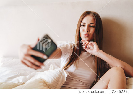 Woman taking a selfie while sitting on bed with white sheets in a cozy indoor setting during daytime Woman taking a selfie while sitting on bed with white sheets in a cozy indoor setting during daytime 128362233