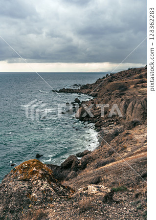 Coastal landscape under cloudy skies with rocky shoreline and gentle waves along the shore 128362283