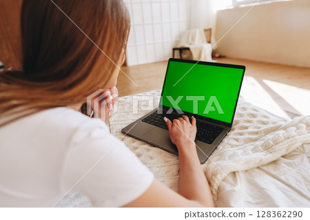 Young person using a laptop with a green screen while sitting on a bed in a cozy room during the day 128362290