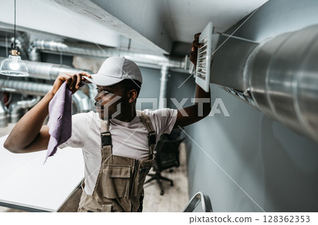 Maintenance worker performs air vent cleaning in modern office setting to ensure optimal air quality and comfort for employees during work hours Maintenance worker performs air vent cleaning in modern office setting to ensure optimal air quality and comfort for employees during work hours 128362353