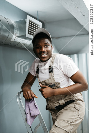 Worker smiling while on ladder, preparing for home maintenance in a cozy indoor space during daylight hours Worker smiling while on ladder, preparing for home maintenance in a cozy indoor space during daylight hours 128362357