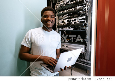 Young professional managing servers with laptop while smiling in a data center during daytime 128362358