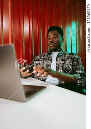 Focused young man working on a laptop while using a smartphone in a vibrant workspace 128362359