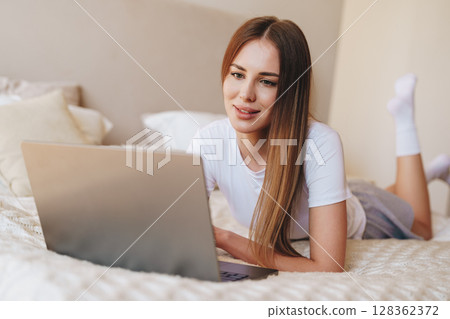 Young woman relaxing and working on laptop while lying on bed in cozy bedroom setting 128362372