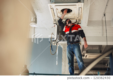 Construction worker performing maintenance on ceiling equipment in modern workspace during daylight hours 128362430