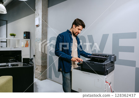 Man operates a printer in a modern office environment during daylight, focused on completing tasks efficiently and professionally 128362437