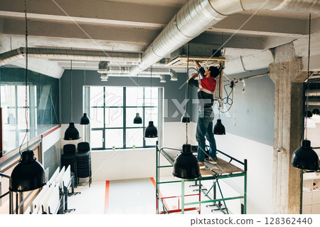 Construction worker performing maintenance on ceiling equipment in modern workspace during daylight hours Construction worker performing maintenance on ceiling equipment in modern workspace during daylight hours 128362440