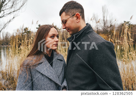 Couple sharing an intimate moment by the lake surrounded by tall grass on a cloudy day 128362505