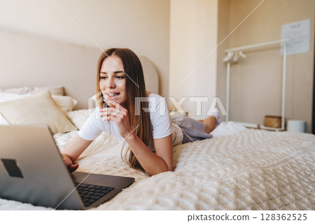 Young woman working on laptop while relaxing on bed in a cozy bedroom during daytime 128362525