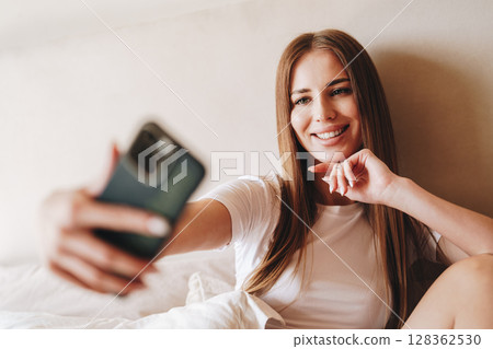 Woman taking a selfie on her smartphone while sitting on a bed in a cozy room during daylight Woman taking a selfie on her smartphone while sitting on a bed in a cozy room during daylight 128362530