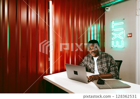 Man working on laptop at desk in modern workspace with neon exit sign illuminating the background Man working on laptop at desk in modern workspace with neon exit sign illuminating the background 128362531
