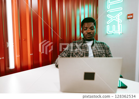 Young man working on laptop in modern workspace with vibrant orange and green decor 128362534