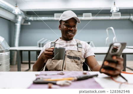 Man in work attire enjoys coffee while looking at phone in modern workspace during afternoon hours 128362546