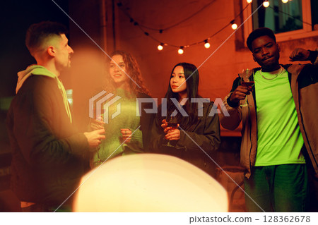 Friends gather around a warm outdoor gathering enjoying drinks and laughter at night under string lights 128362678