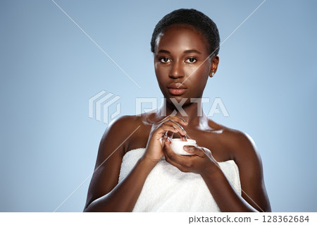 Woman applying cream while wearing a towel, capturing essence of skincare routine against a blue background Woman applying cream while wearing a towel, capturing essence of skincare routine against a blue background 128362684