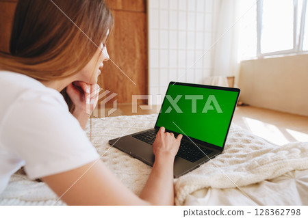Young woman using laptop while lounging on bed in bright indoor space during daytime Young woman using laptop while lounging on bed in bright indoor space during daytime 128362798