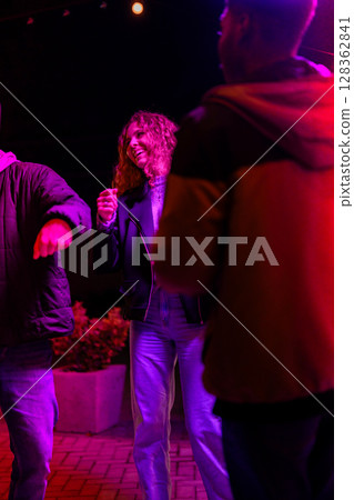 People enjoy dancing and socializing at an outdoor event under colorful lights during the evening 128362841