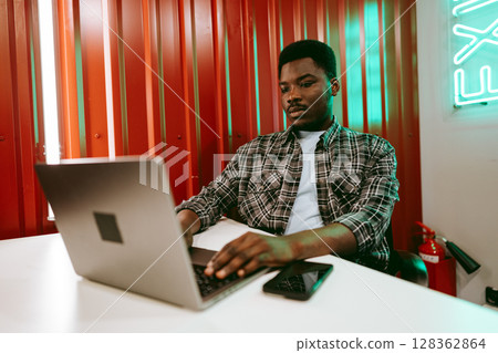 Young man working on laptop in modern workspace with vibrant red walls and neon lighting during daytime hours 128362864