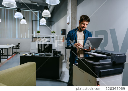 Man using laptop in modern office environment while standing near printer in bright work space Man using laptop in modern office environment while standing near printer in bright work space 128363069