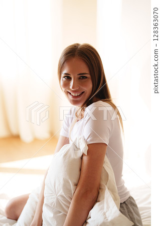 Young woman smiling while holding a pillow in a bright, cozy bedroom during morning light Young woman smiling while holding a pillow in a bright, cozy bedroom during morning light 128363070