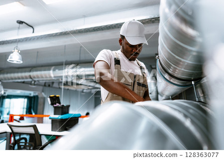 Worker engages in mechanical repairs inside a modern workshop during daylight hours Worker engages in mechanical repairs inside a modern workshop during daylight hours 128363077