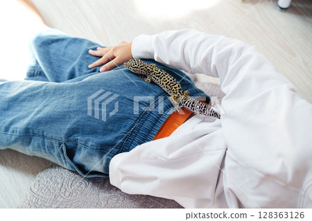 Child relaxed on a rug with a gecko exploring their waistband indoors in a bright room 128363126
