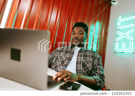 Young man working on laptop in a modern workspace with vibrant red walls and bright exit sign at evening 128363201