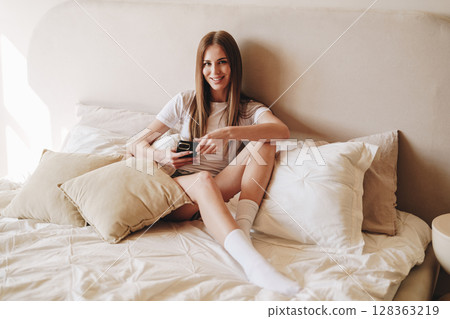 Smiling woman relaxing on a cozy bed while using her phone in a well-lit room during the day Smiling woman relaxing on a cozy bed while using her phone in a well-lit room during the day 128363219