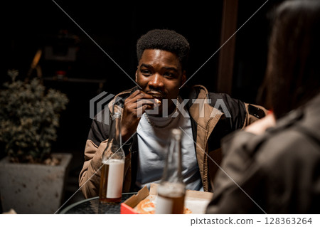Young man enjoying snack outdoors at night with friends while having drinks and conversing Young man enjoying snack outdoors at night with friends while having drinks and conversing 128363264