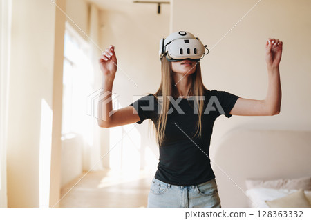 Woman experiencing virtual reality in a bright room during daylight hours while engaging in an interactive digital environment Woman experiencing virtual reality in a bright room during daylight hours while engaging in an interactive digital environment 128363332
