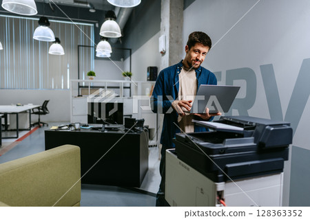 Man using a laptop while standing next to a printer in a modern office setting during daytime 128363352