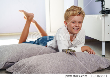 Young boy enjoying time at home while playing with a pet gecko on soft pillows in a bright room 128363361