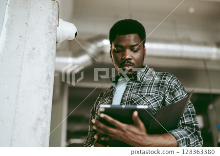 Young man reviewing documents on tablet while standing in modern workspace with industrial design elements 128363380