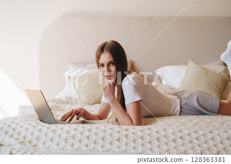 Young woman working on laptop while relaxing on bed with pillows during daytime in cozy room Young woman working on laptop while relaxing on bed with pillows during daytime in cozy room 128363381