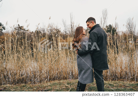 Couple enjoys a romantic moment by a serene waterway during autumn in a park during cool weather Couple enjoys a romantic moment by a serene waterway during autumn in a park during cool weather 128363407