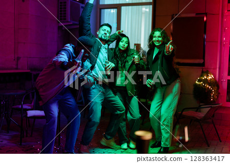 Friends celebrate joyfully at night with colorful lights in a cozy outdoor gathering Friends celebrate joyfully at night with colorful lights in a cozy outdoor gathering 128363417