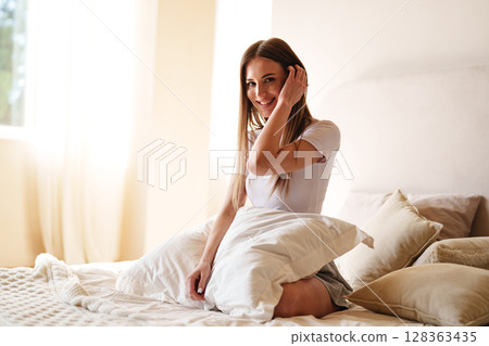 Smiling woman sitting on bed with pillows in a sunlit room during morning 128363435