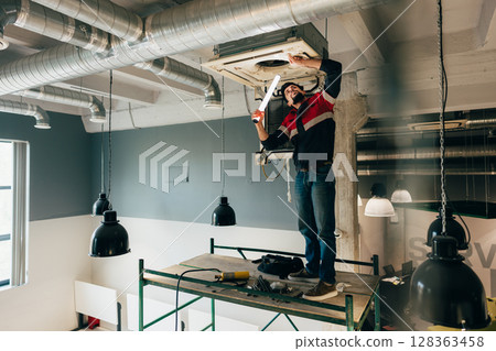 Worker repairs air conditioning unit while standing on scaffold in modern workspace during daylight hours Worker repairs air conditioning unit while standing on scaffold in modern workspace during daylight hours 128363458