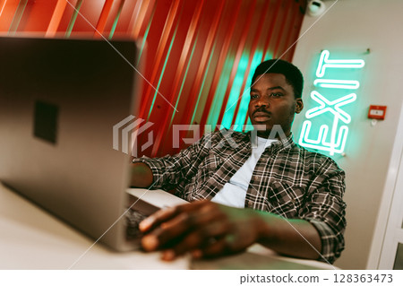 Young man working on laptop in modern workspace with neon exit sign on red wall 128363473